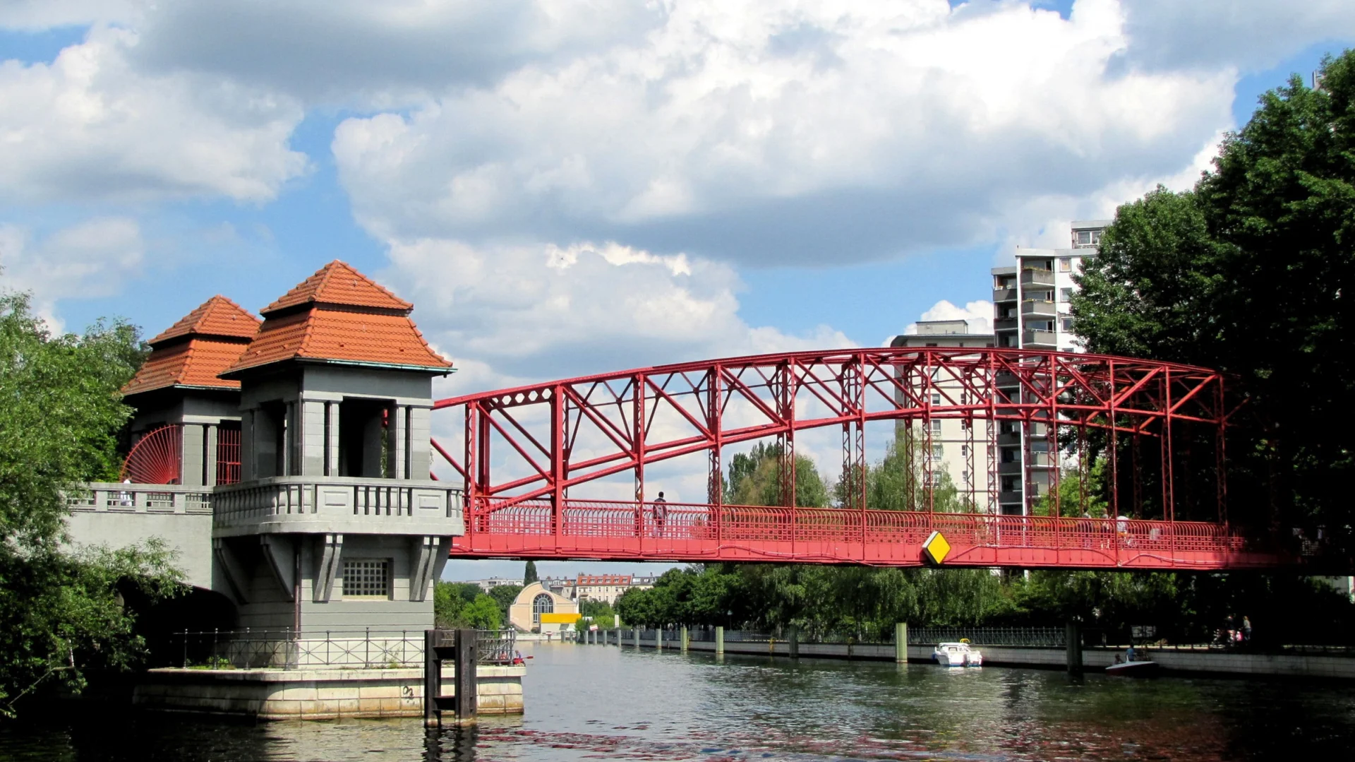 Rote Eisenbrücke mit Türmen und roten Dächern über einem Fluss, im Hintergrund Wohnhäuser.