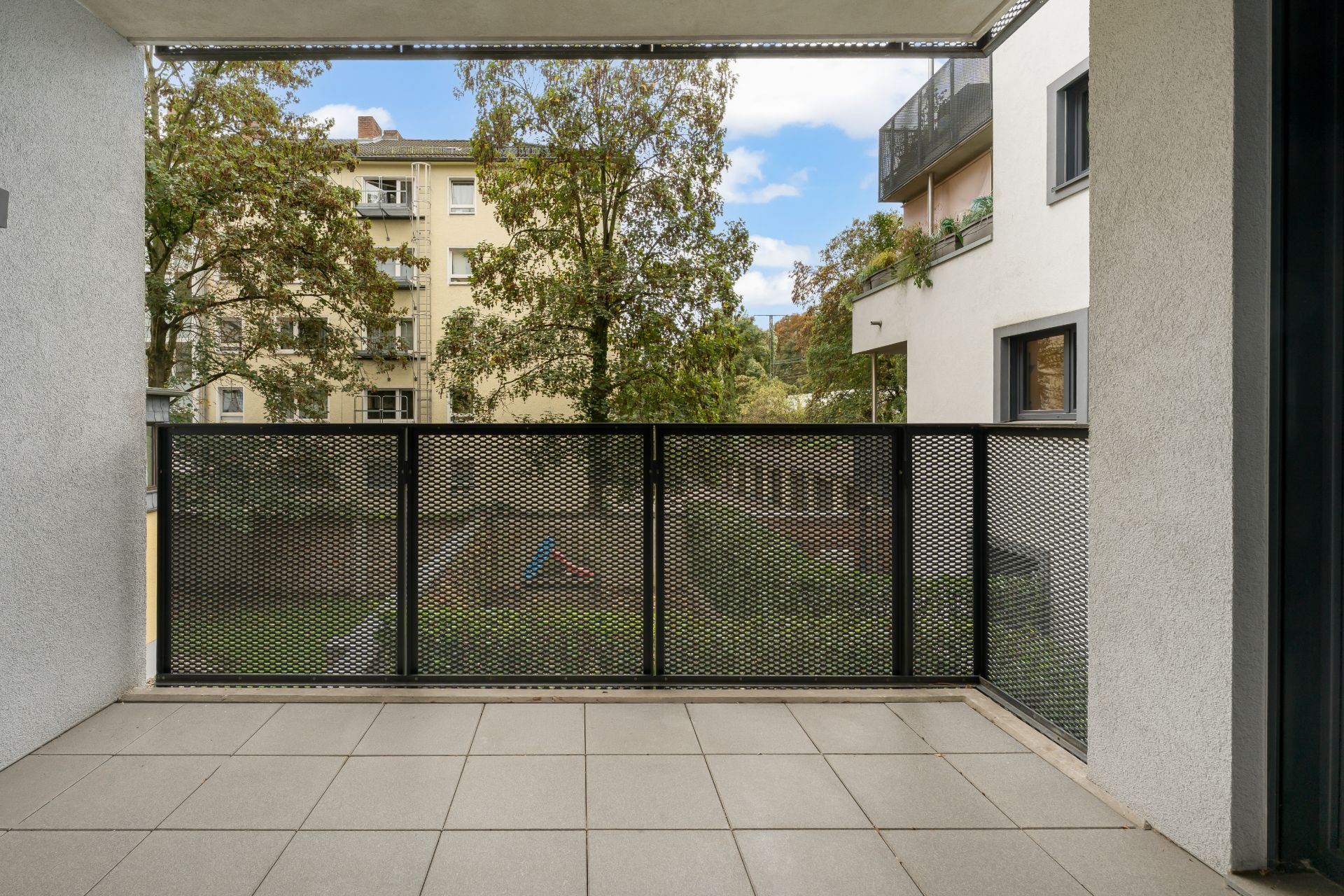 Balkon mit Fliesenboden, Metallgeländer und Blick auf Bäume sowie angrenzende Wohngebäude.