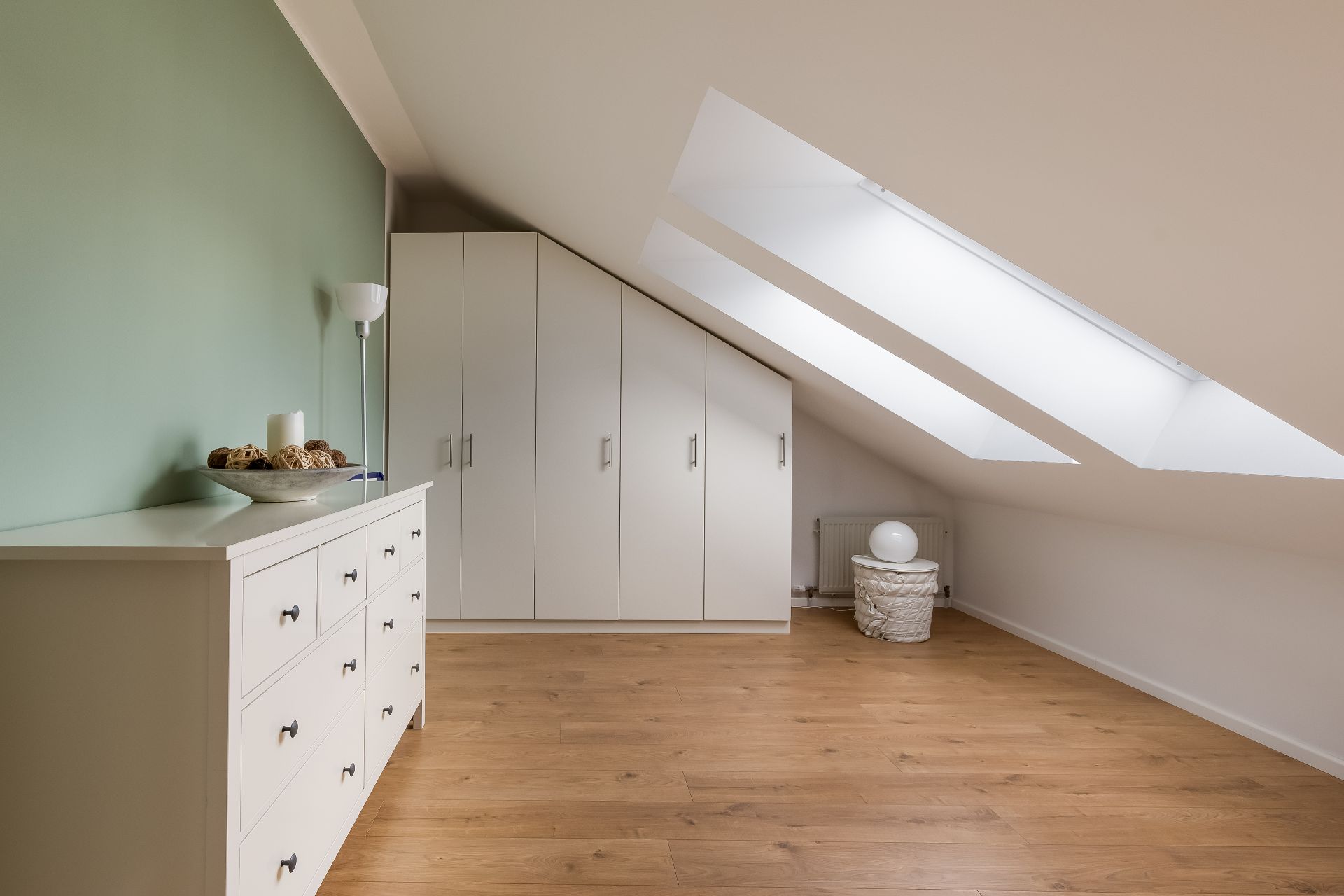 Heller Dachraum mit Schrägfenstern, Kommode, Einbauschrank und Holzfußboden.
