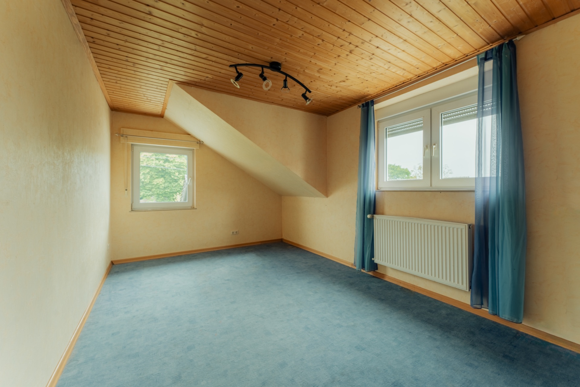 Leeres Zimmer mit schräger Decke, zwei Fenstern, blauem Teppich und Holzdecke.