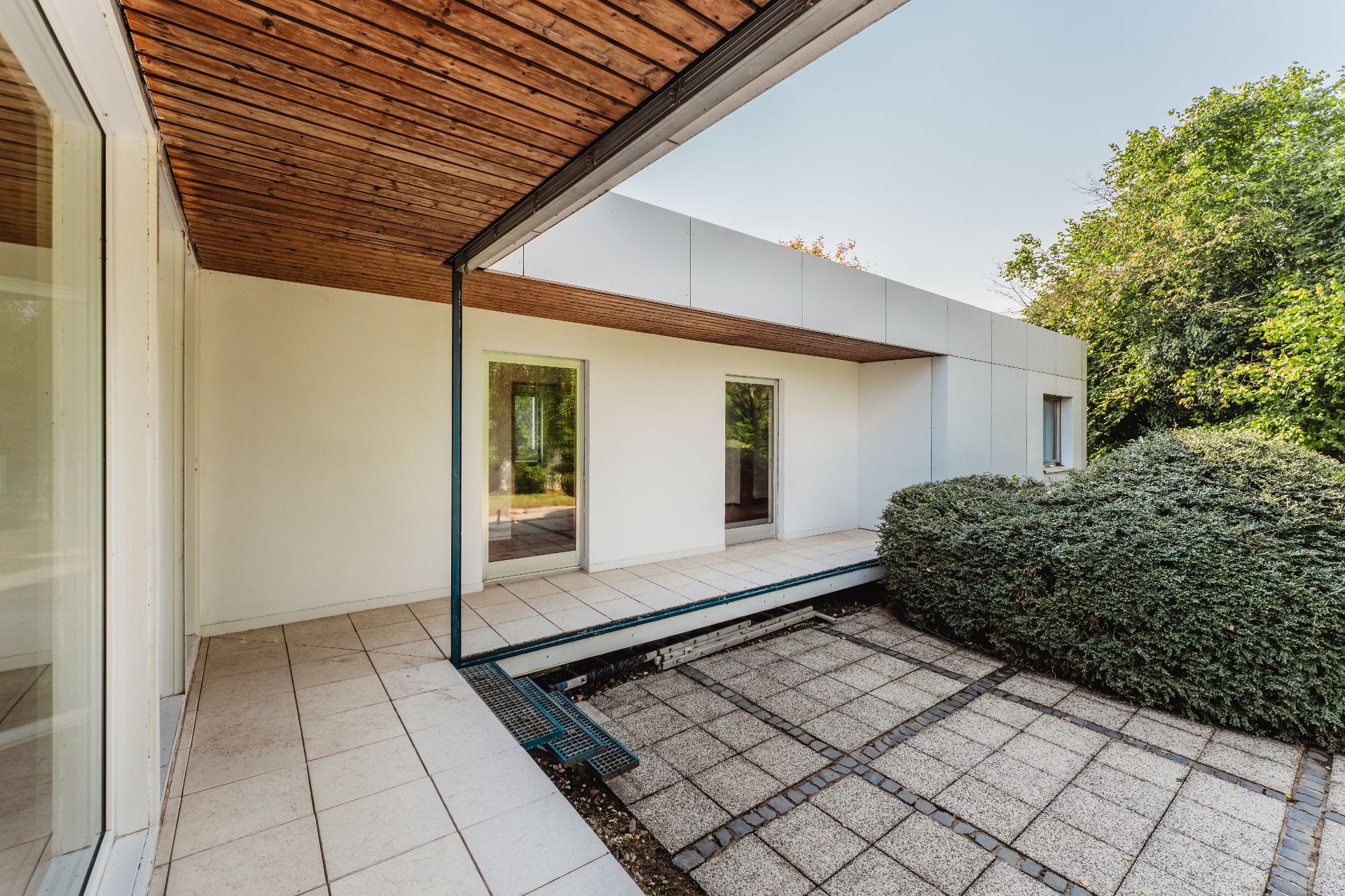 Moderne Terrasse mit Holzdach, weißen Wänden und Fliesen neben grünen Büschen.