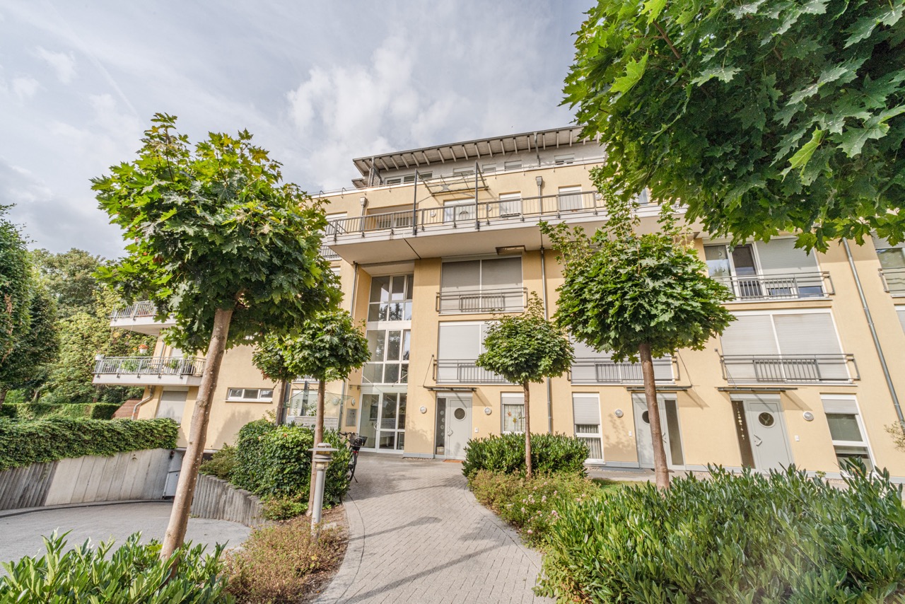 Mehrstöckiges Wohnhaus mit Balkonen, großen Fenstern und gepflegtem Weg, umgeben von Bäumen und Sträuchern.