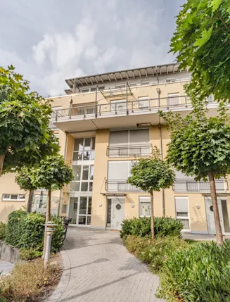 Mehrstöckiges Wohnhaus mit Balkonen, großen Fenstern und gepflegtem Weg, umgeben von Bäumen und Sträuchern.