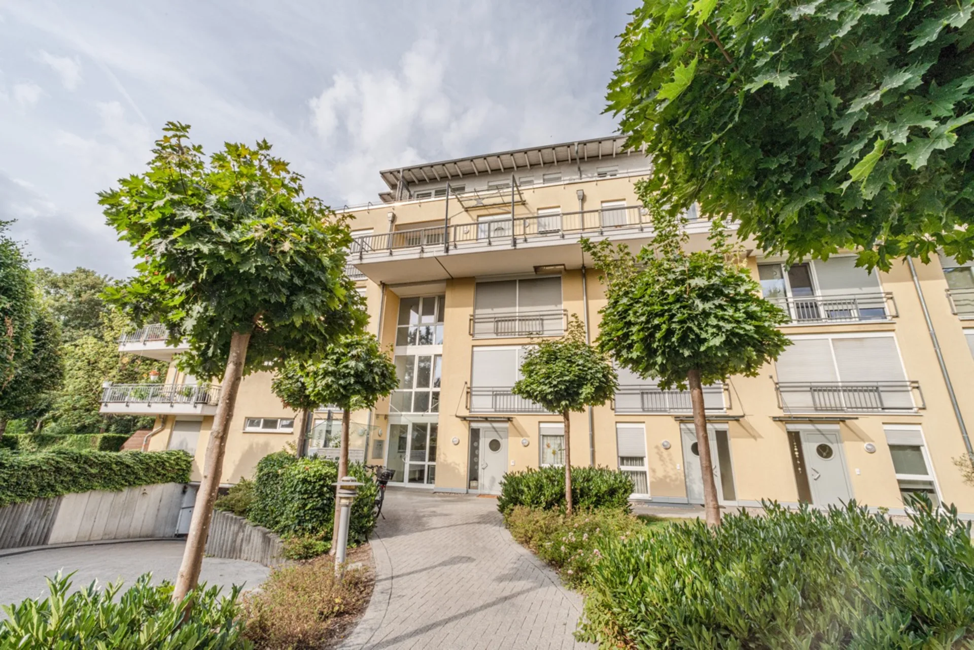 Mehrstöckiges Wohnhaus mit Balkonen, großen Fenstern und gepflegtem Weg, umgeben von Bäumen und Sträuchern.