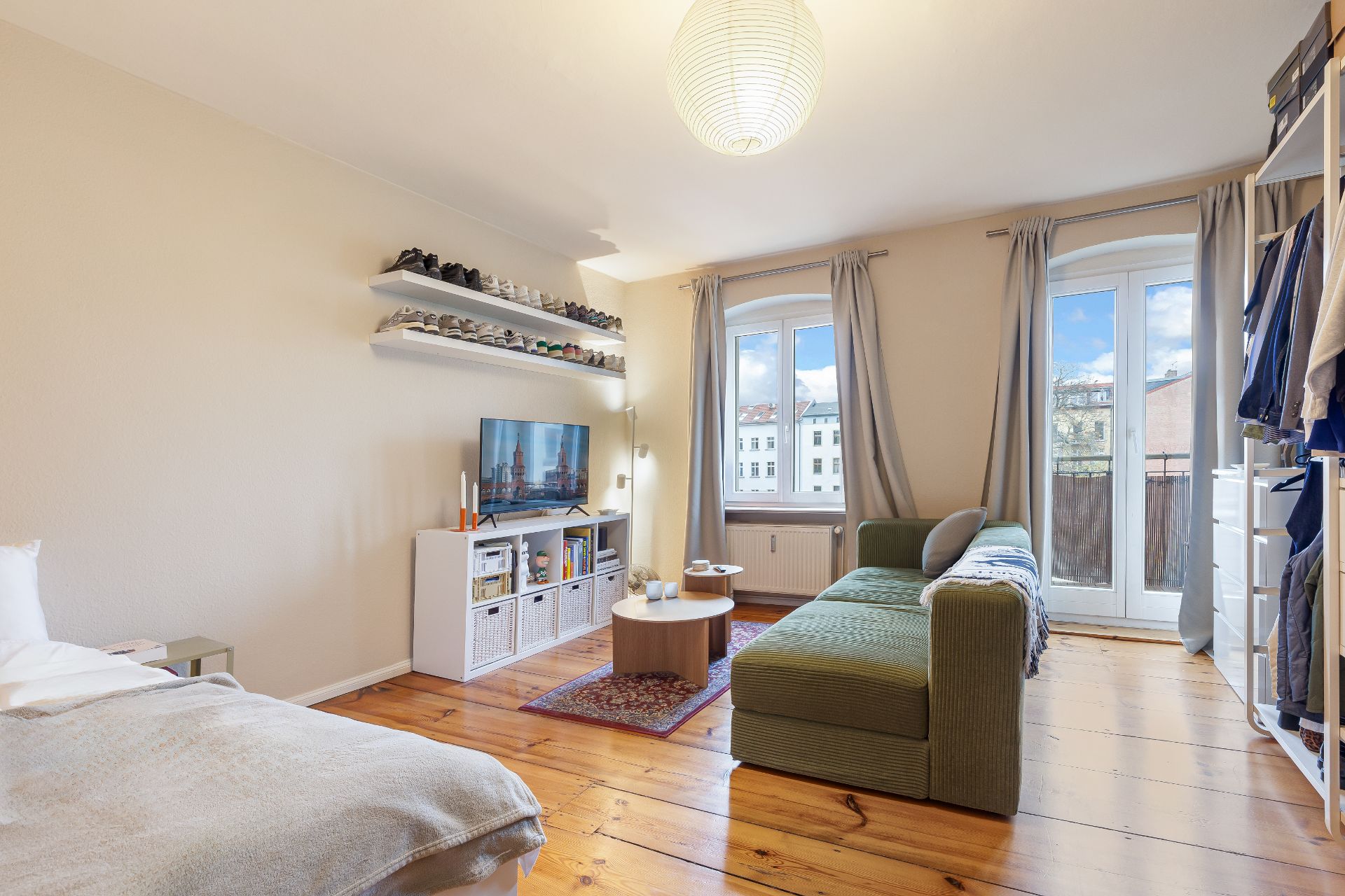 Helles Wohnzimmer mit grünem Sofa, TV, Regal, Bett und Holzboden, zwei Fenstern und Balkon.