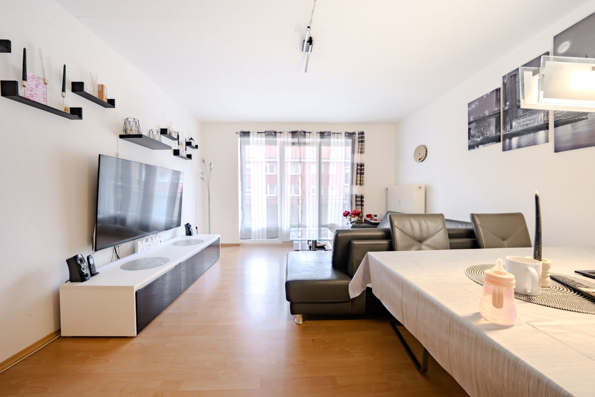Helles Wohnzimmer mit Fernseher, Sofa, Esstisch und großen Fenstern mit blickdichten Vorhängen.