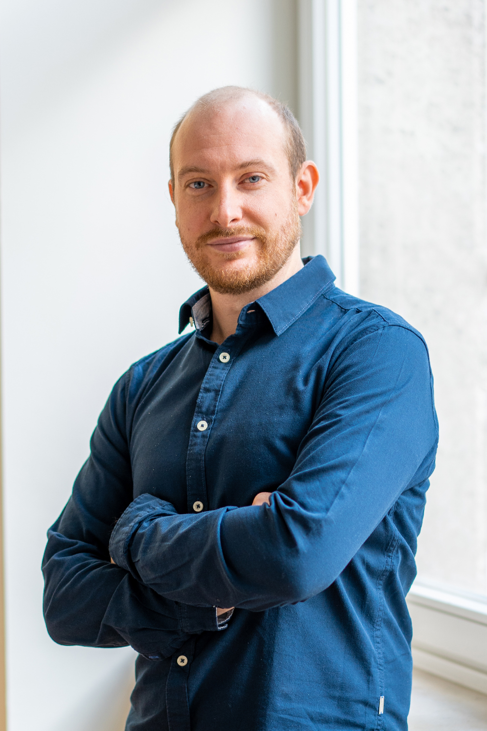 Eine Person mit blauem Hemd steht mit verschränkten Armen vor einem Fenster.