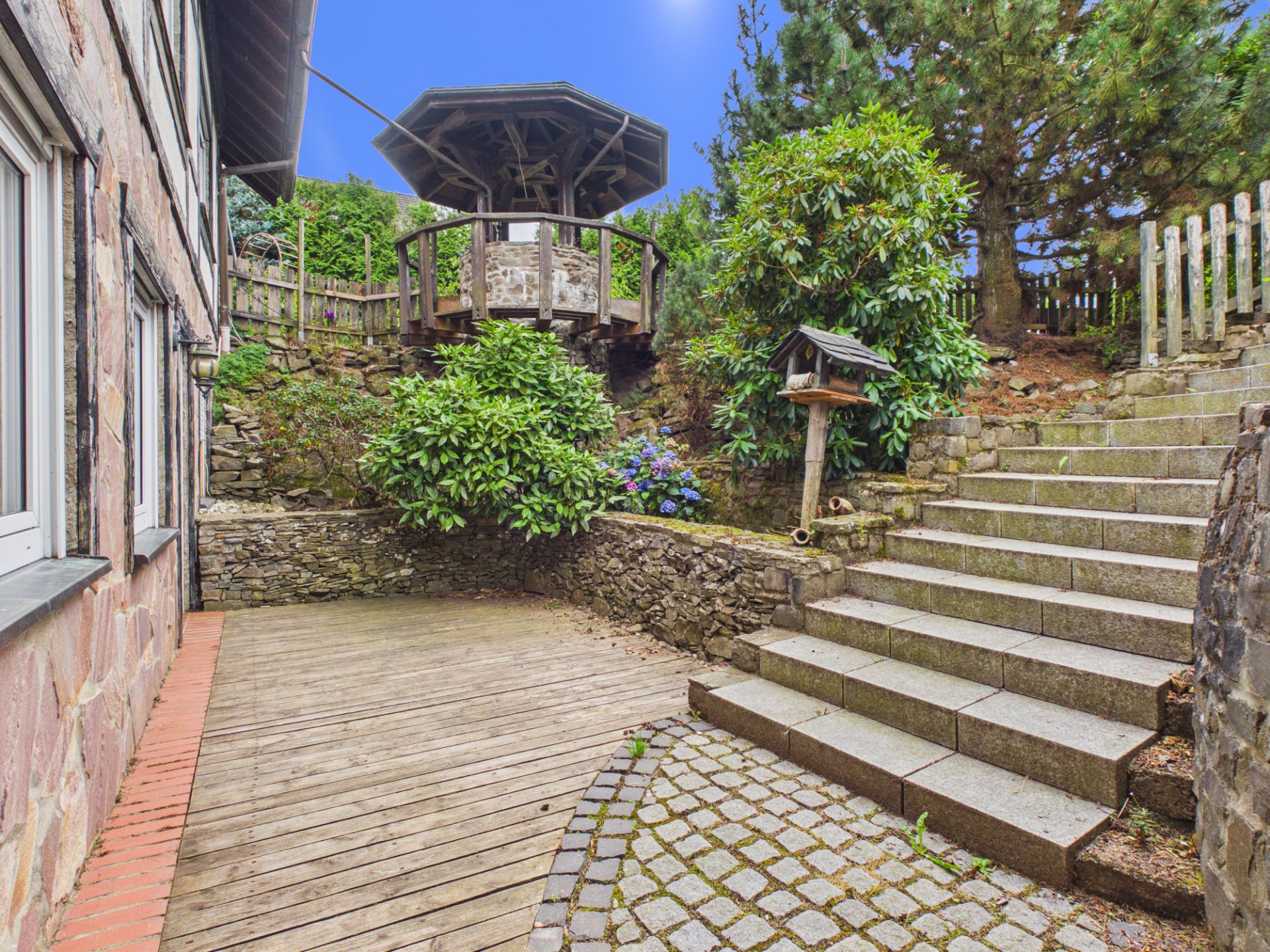 Gepflasterter Hof mit Treppe, Holzterrasse, Pflanzen und einem überdachten Brunnen im Garten.