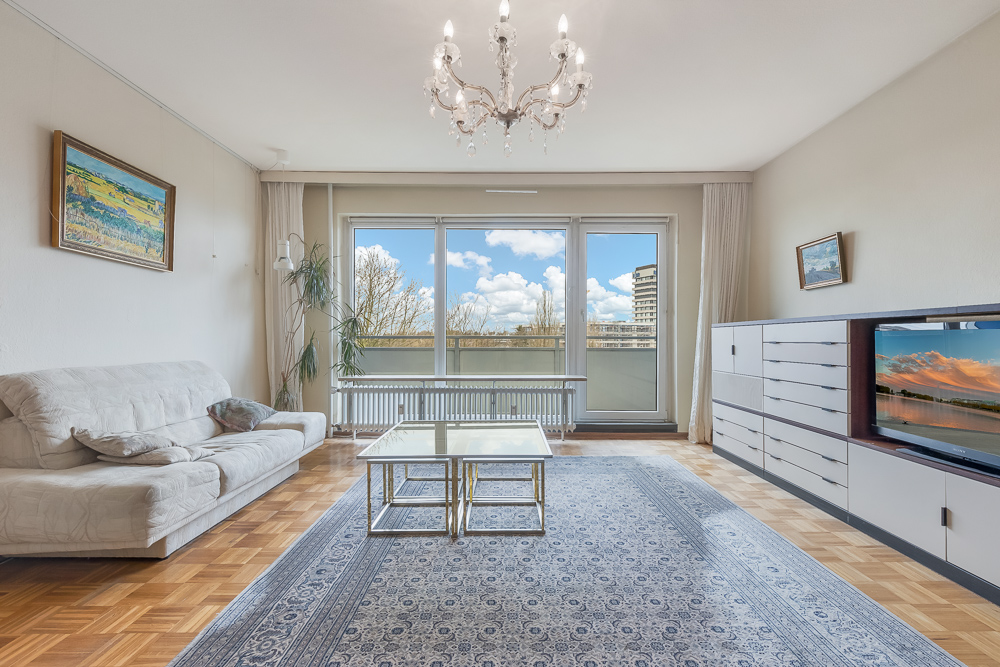 Helles Wohnzimmer mit Sofa, Teppich, Couchtisch, TV, Kommode und großem Fenster zum Balkon.