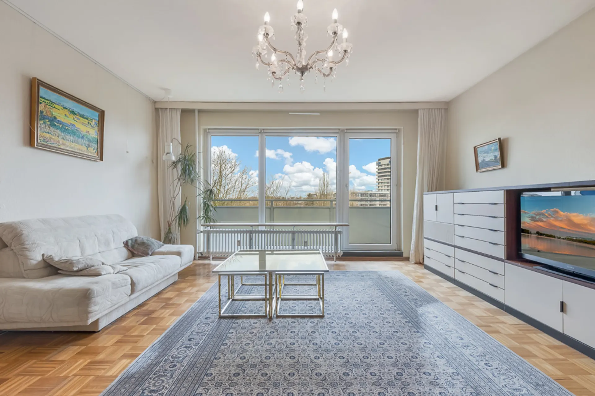 Helles Wohnzimmer mit Sofa, Teppich, Couchtisch, TV, Kommode und großem Fenster zum Balkon.