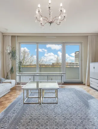 Helles Wohnzimmer mit Sofa, Teppich, Couchtisch, TV, Kommode und großem Fenster zum Balkon.