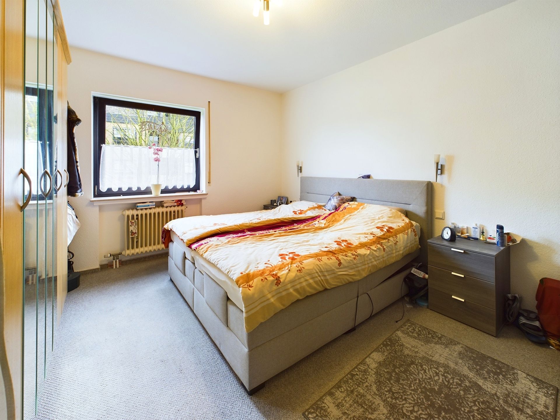 Helles Schlafzimmer mit Doppelbett, Nachttisch, Schrank und Fenster mit Gardinen.