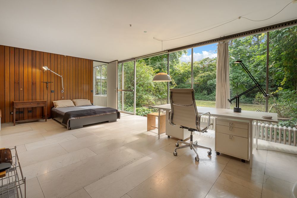 Helles Schlafzimmer mit Holzwand, Bett und großem Schreibtisch vor bodentiefen Fenstern mit Gartenblick.