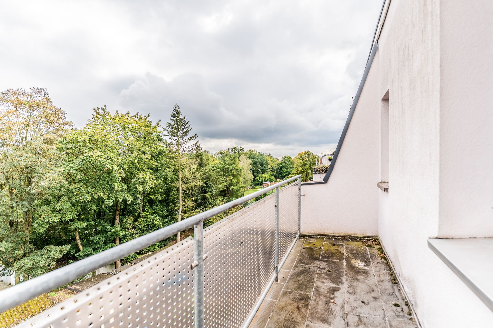 Balkon mit Metallgeländer und Blick auf grüne Bäume unter bewölktem Himmel.