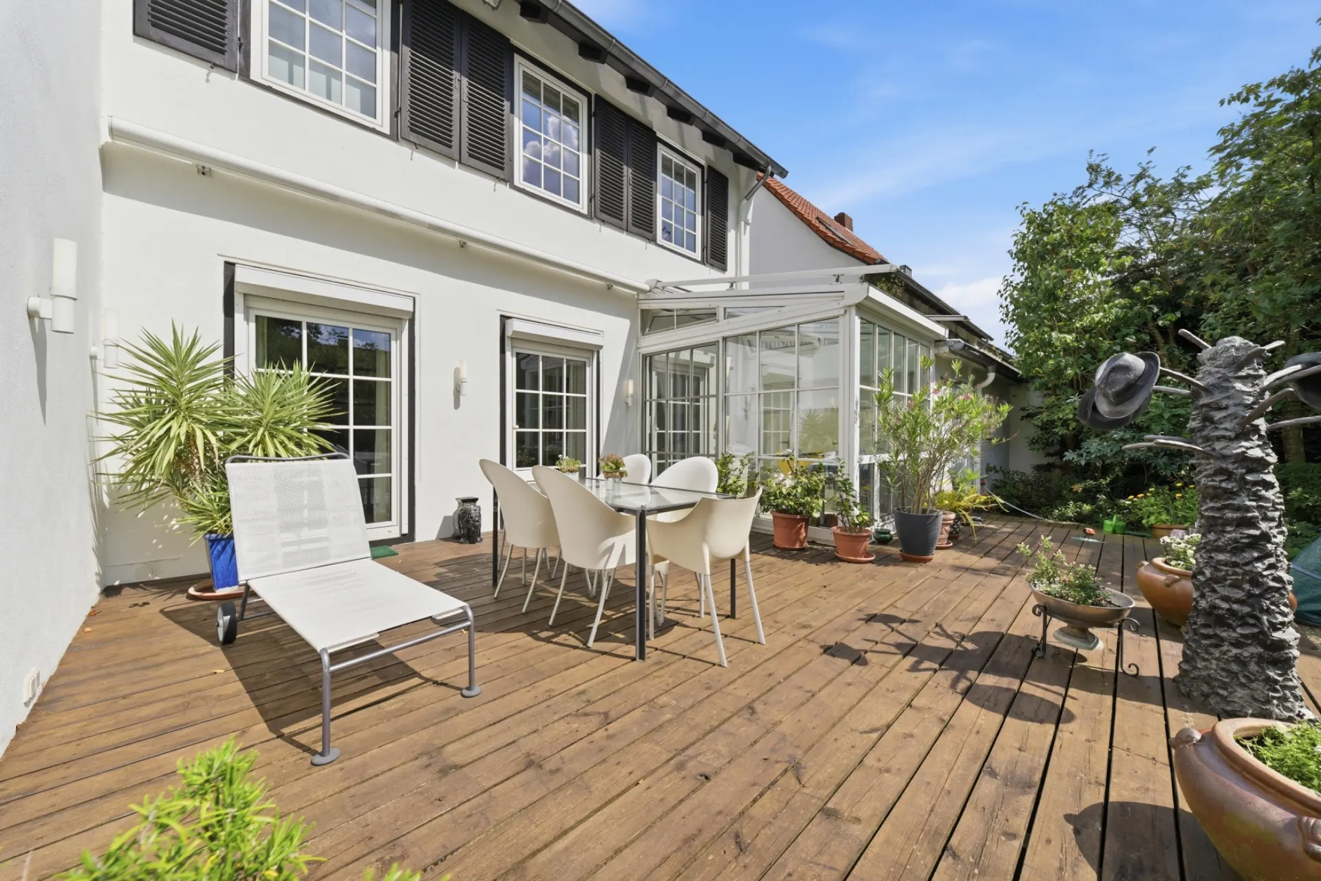 Große Holzterrasse mit Esstisch, Stühlen, Sonnenliege und vielen Pflanzen an einem Wohnhaus.