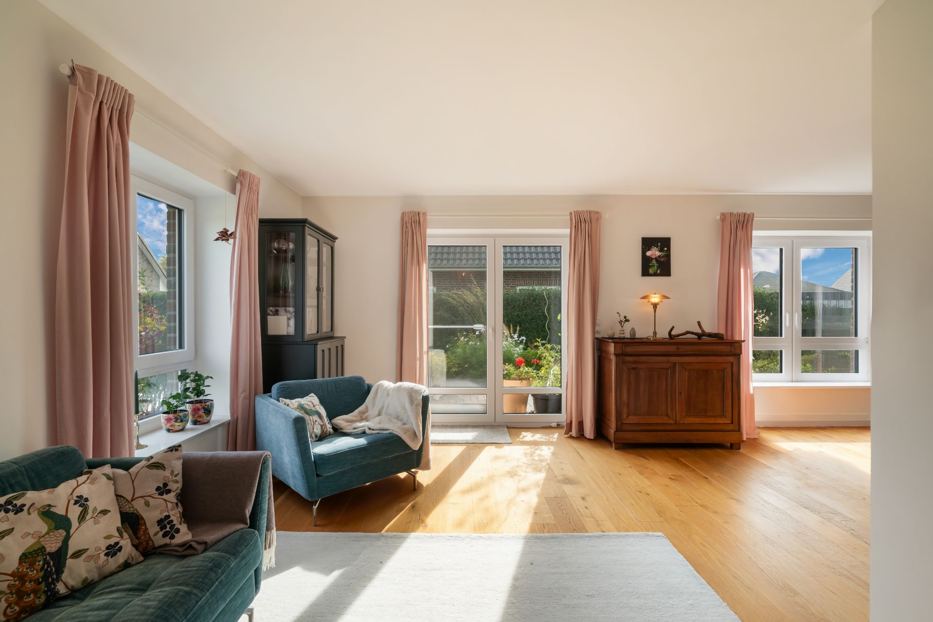Helles Wohnzimmer mit Holzboden, zwei Sofas, Kommode, Fenstern und rosa Vorhängen.