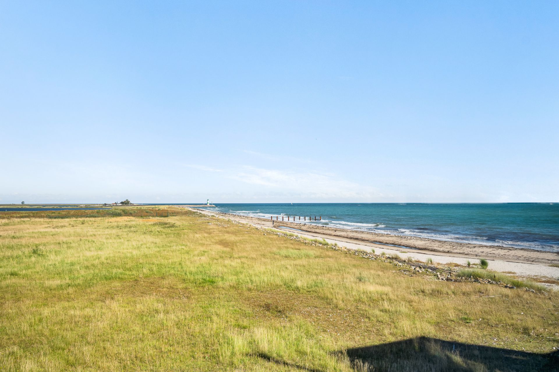 Blick auf eine Wiese mit angrenzendem Sandstrand und Meer unter blauem Himmel.