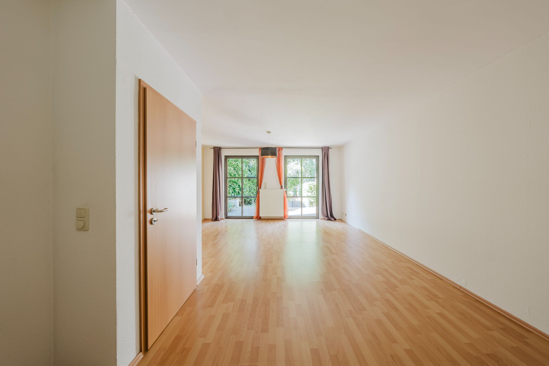 Leeres Zimmer mit Holzfußboden, offener Tür und großen Fenstern mit Blick ins Grüne.