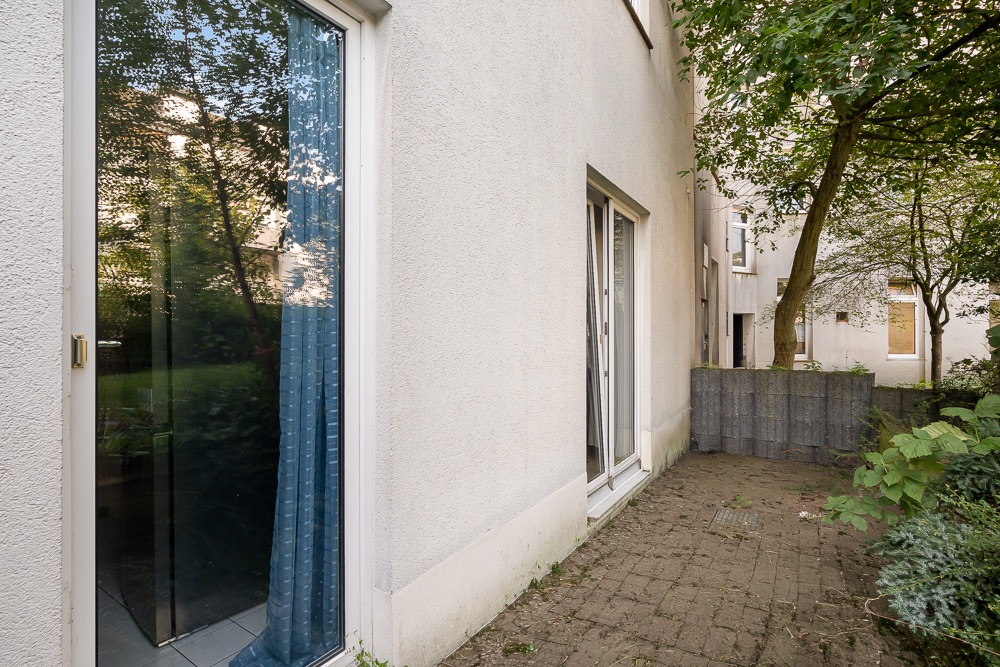 Gepflasterte Terrasse neben einem Wohnhaus mit Glastüren und Bäumen.
