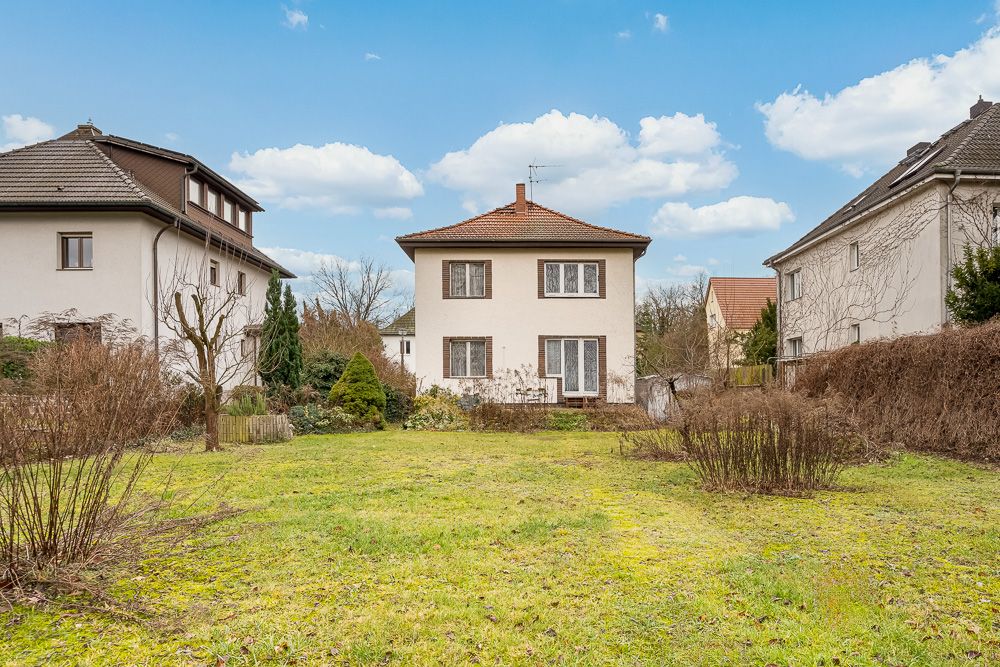 Zweistöckiges Wohnhaus mit Garten, umgeben von weiteren Häusern und Sträuchern bei blauem Himmel.