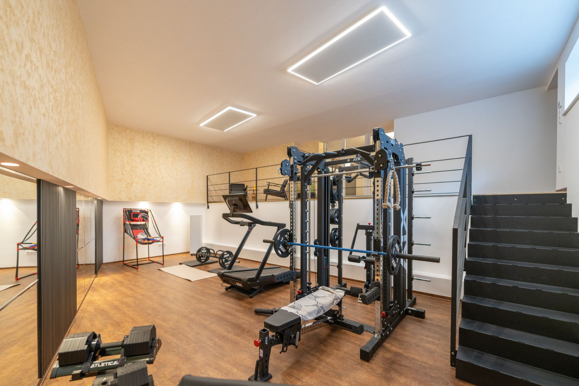 Fitnessraum mit Trainingsgeräten, Laufband und Hantelbank, daneben eine Treppe und Galerie.
