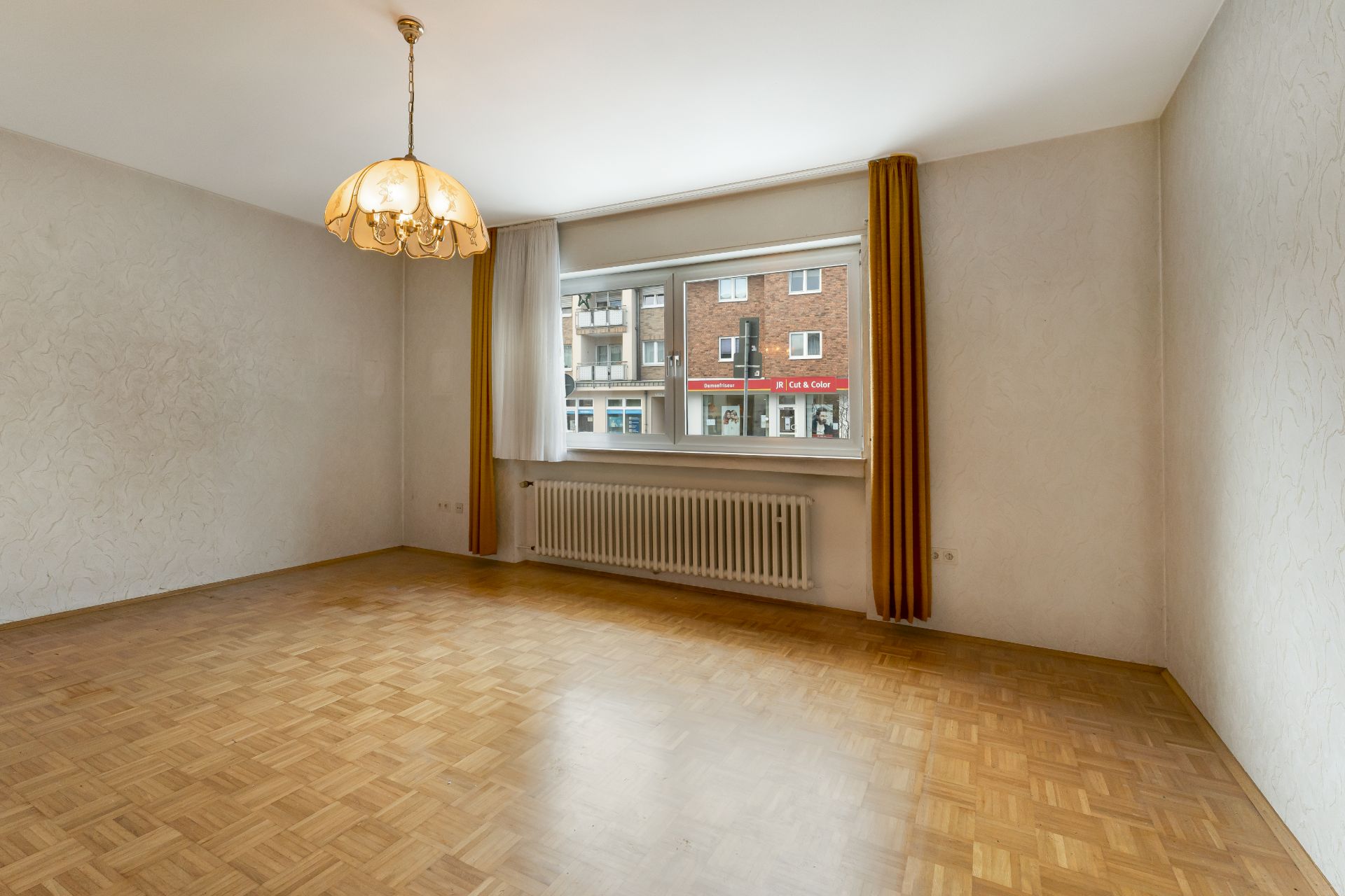 Leeres Zimmer mit Parkettboden, großem Fenster, Vorhängen und Deckenlampe.