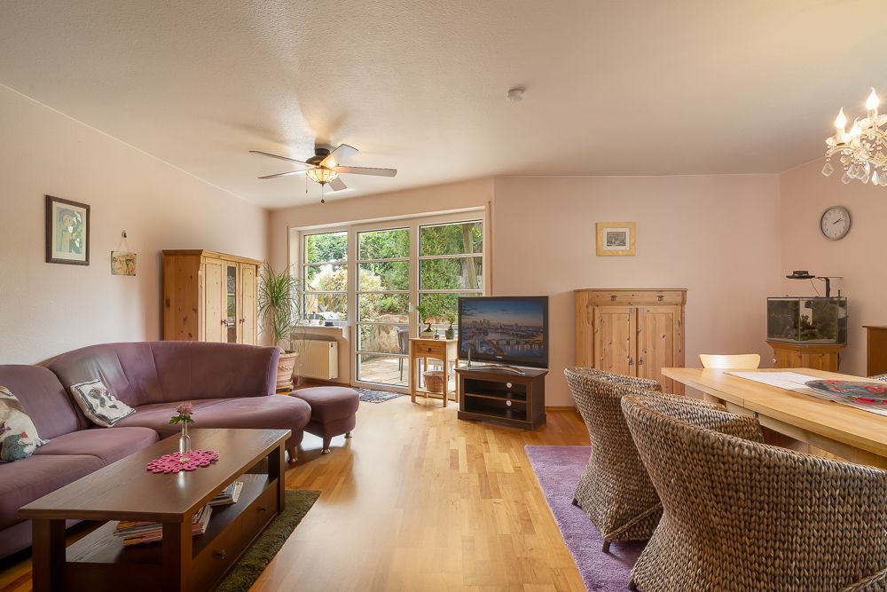 Helles Wohnzimmer mit Sofa, Esstisch, TV, Schrank und großem Fenster zum Garten.