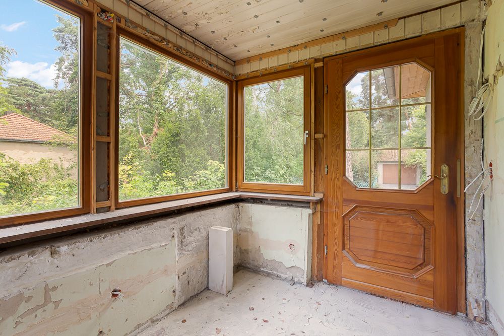 Veranda mit großen Fenstern, Holztür und teils unrenovierten Wänden, Blick ins Grüne.