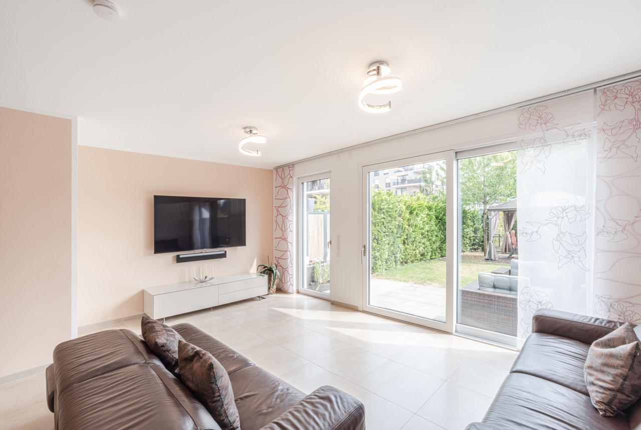 Helles Wohnzimmer mit zwei Sofas, Wand-TV und großen Fenstern mit Blick in den Garten.