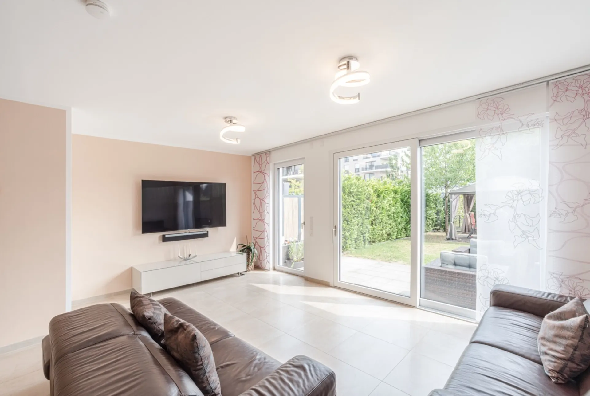 Helles Wohnzimmer mit zwei Sofas, Wand-TV und großen Fenstern mit Blick in den Garten.