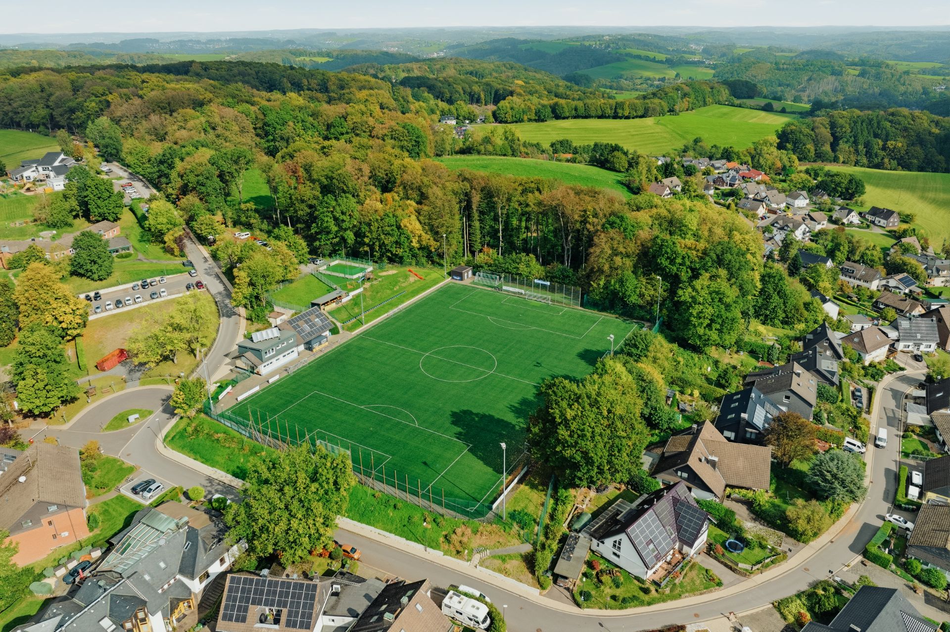Luftaufnahme eines Fußballplatzes inmitten eines Wohngebiets und umgeben von Bäumen.