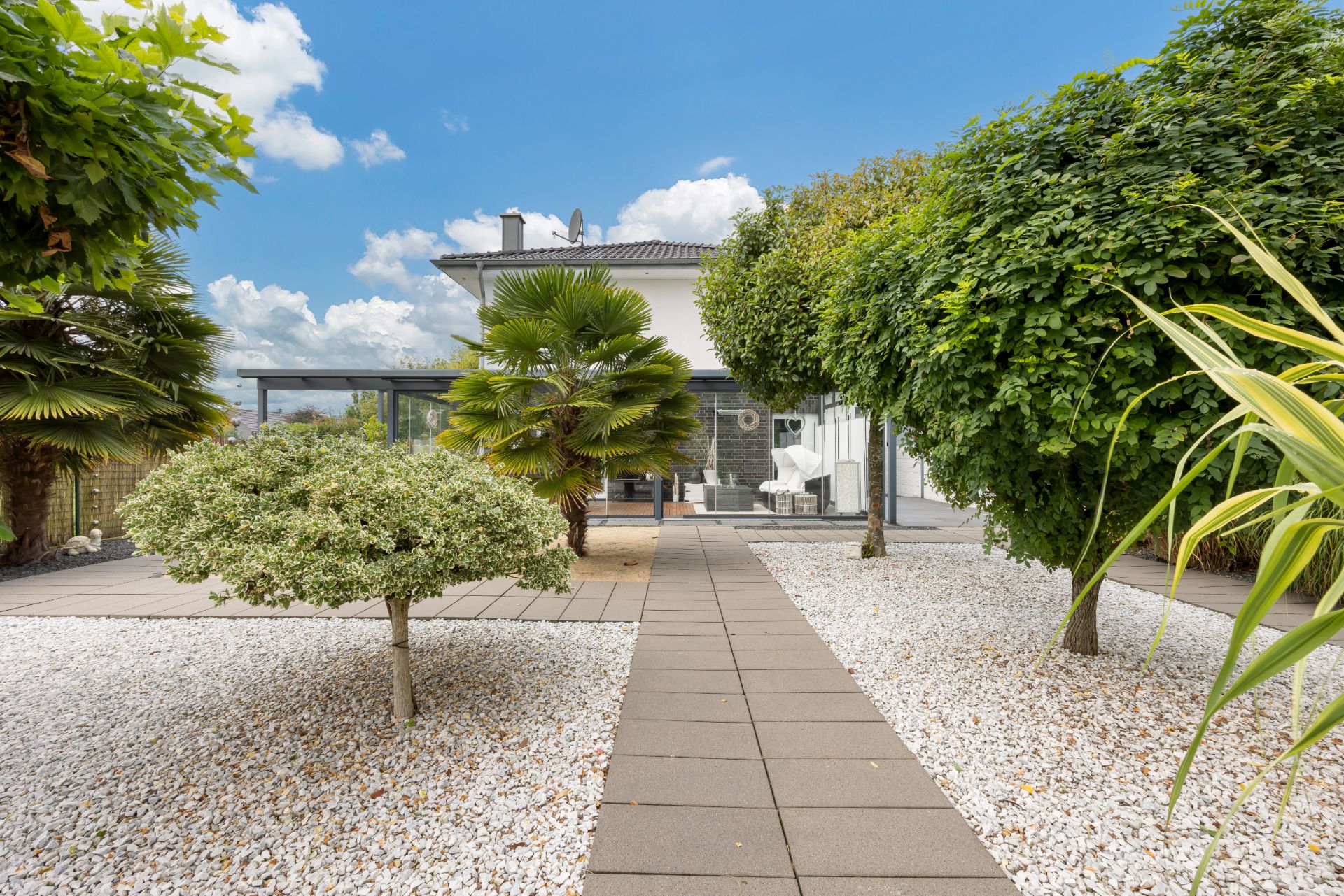 Gepflasterter Weg führt durch einen Garten mit Kies und Bäumen zu einem modernen Wohnhaus.