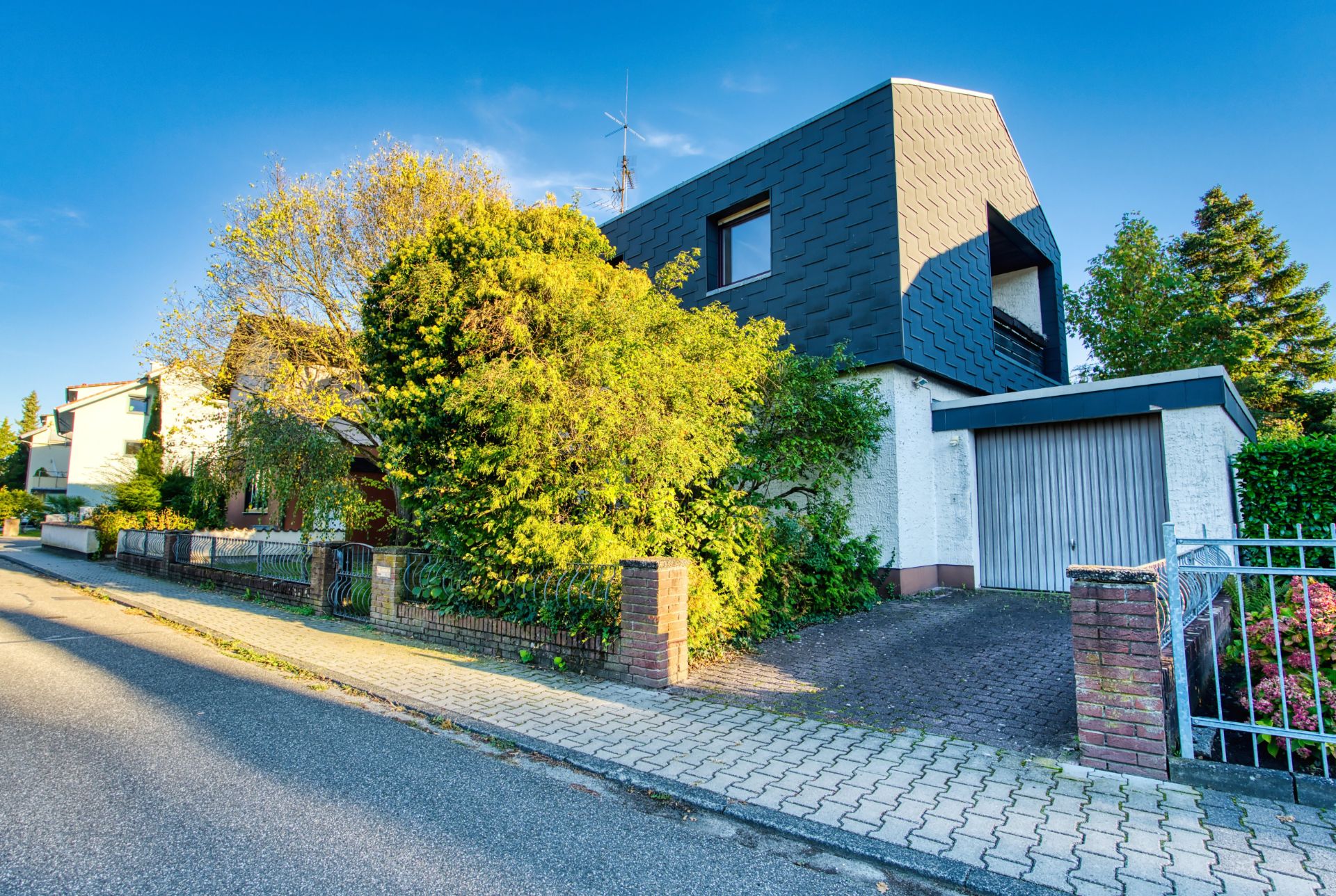 Modernes Wohnhaus mit Garage, Garten und Bäumen an einer ruhigen Wohnstraße.