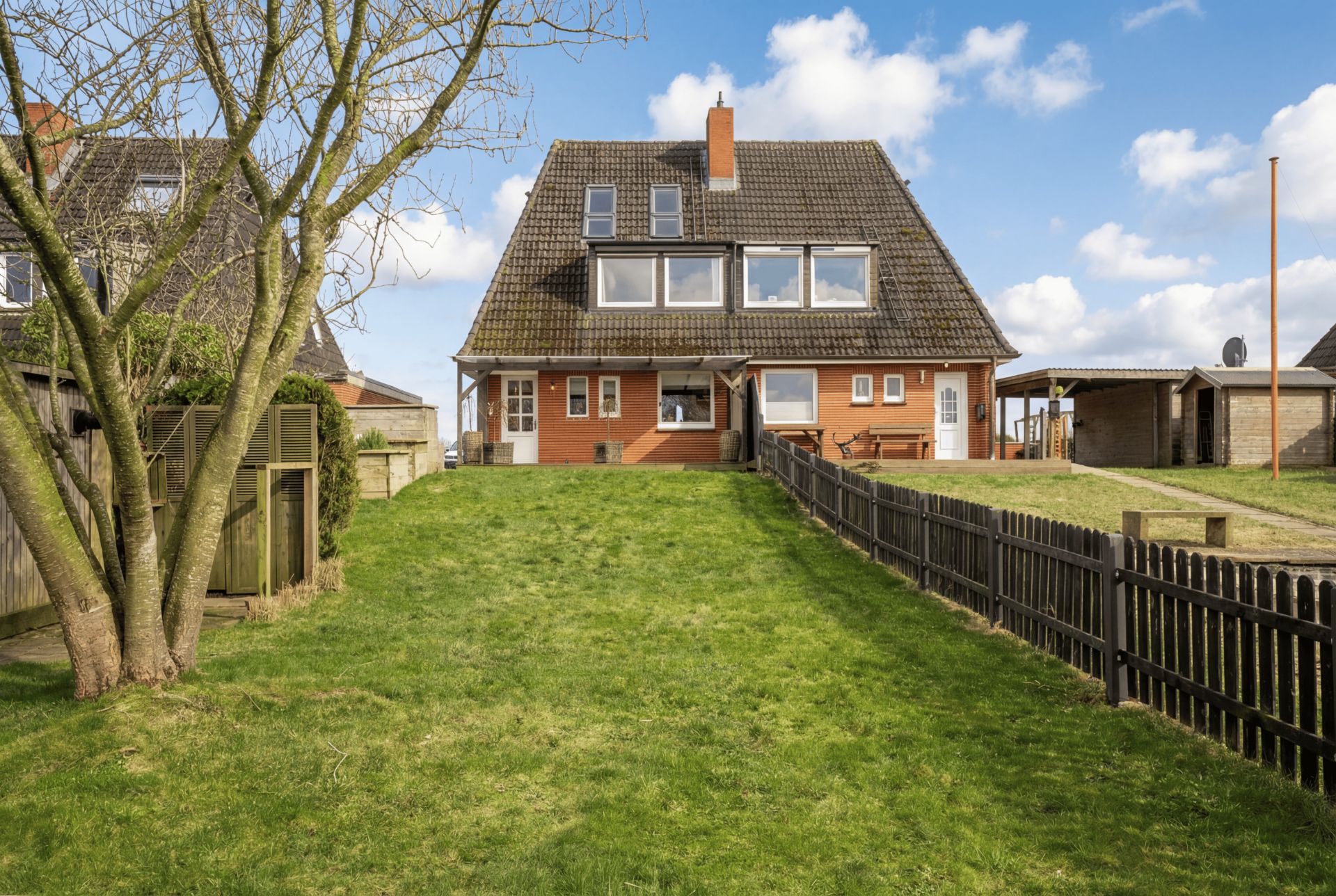 Zweistöckiges Haus mit großem Garten, Holzzaun und Baum im Vordergrund bei Tag.