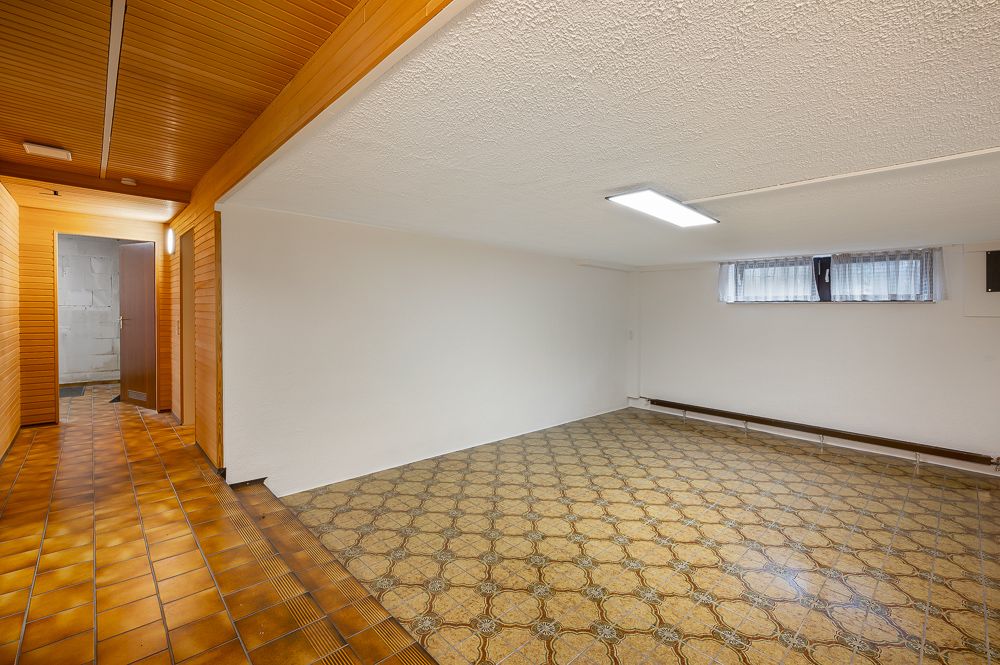 Leerer Kellerraum mit gemustertem Boden, weißer Wand, Holzdecke und kleinem Fenster.