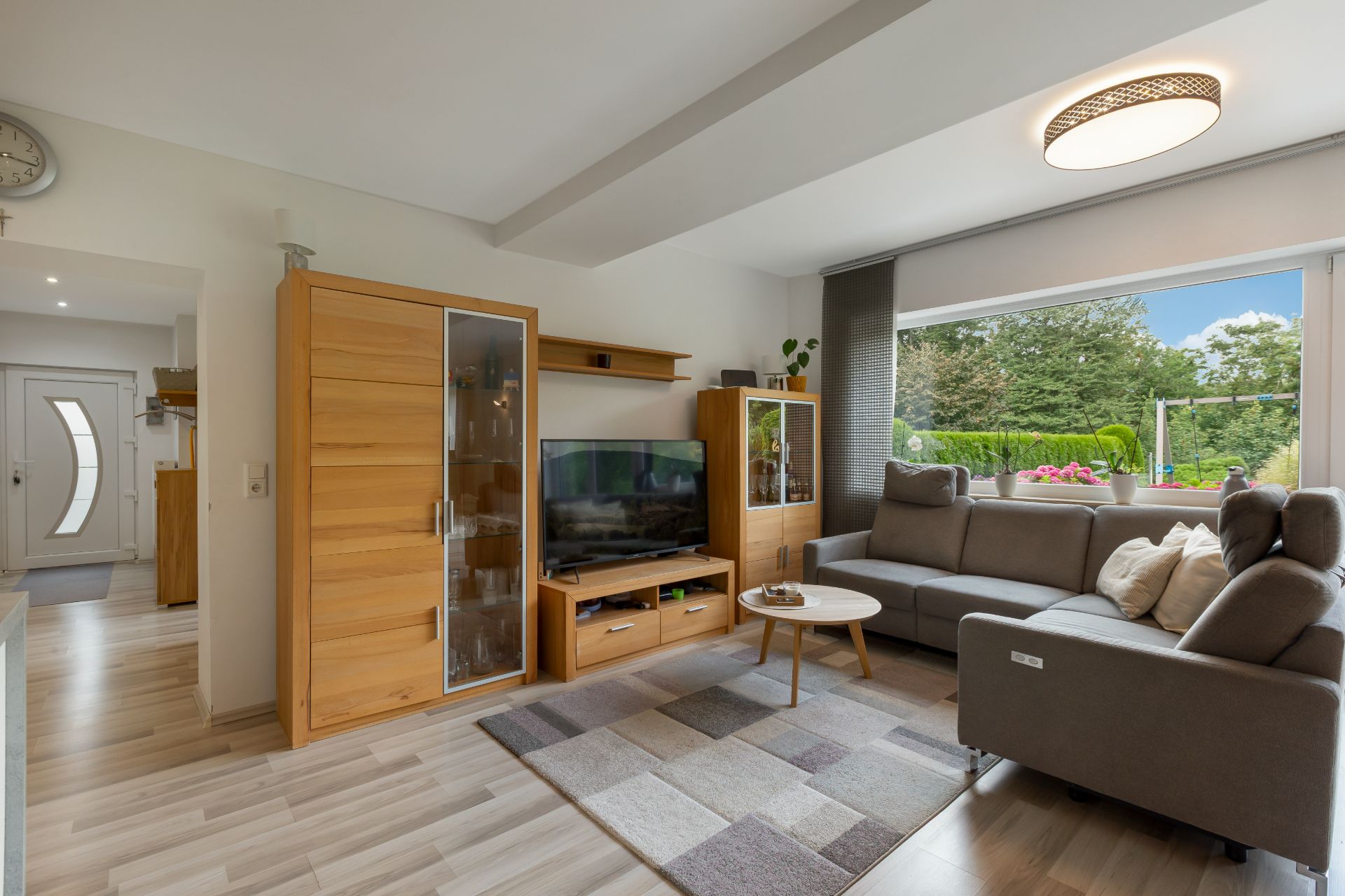 Modernes Wohnzimmer mit Ecksofa, Teppich, TV, Holzschränken und großem Fenster mit Ausblick ins Grüne.
