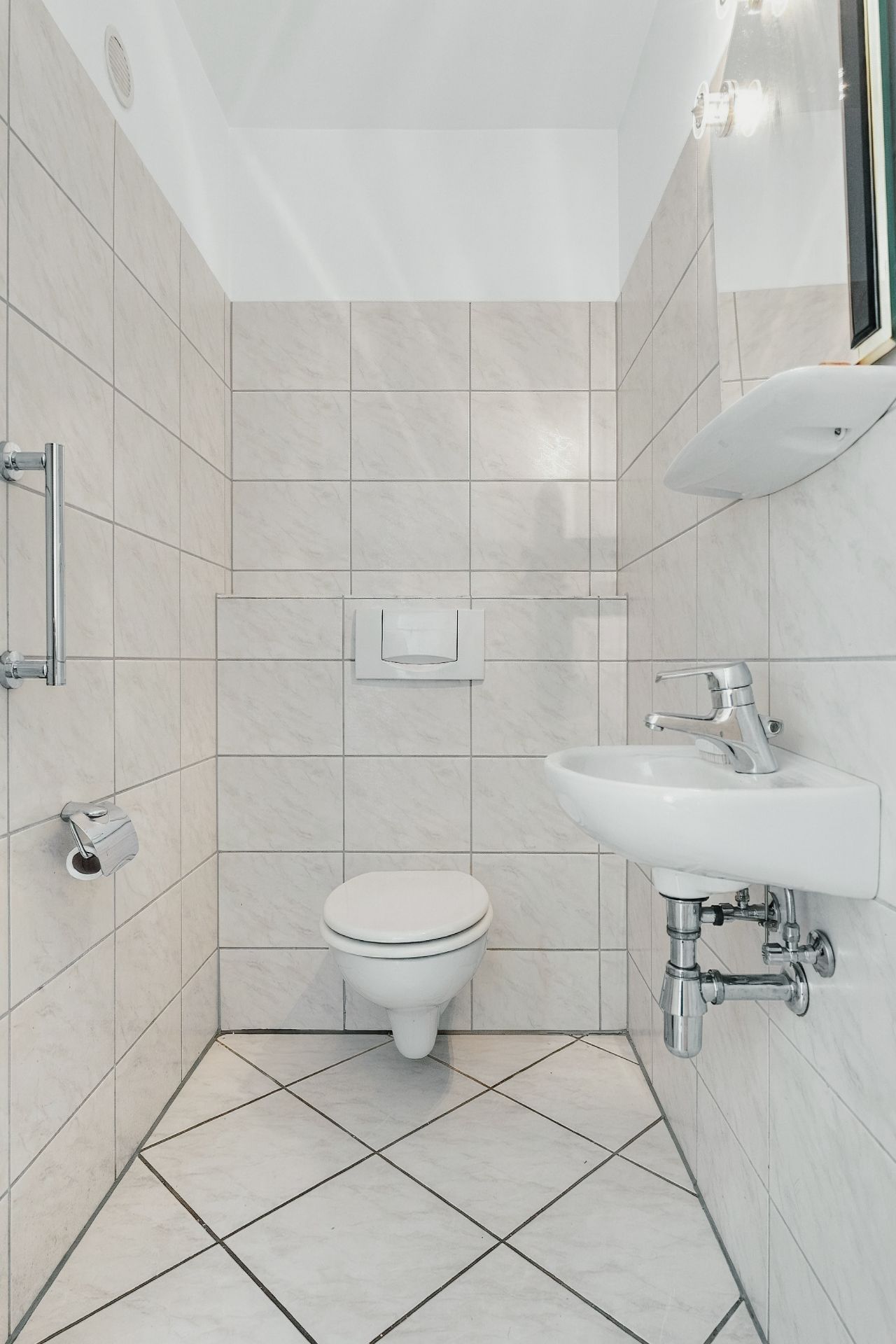 Kleines, gefliestes Bad mit Wandtoilette, Waschbecken und Spiegel.