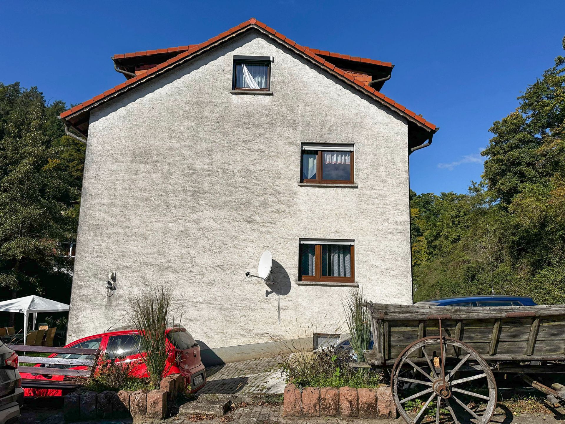 Weiße Hauswand mit drei Fenstern, Satellitenschüssel, rotem Auto und Holzwagen im Vordergrund.