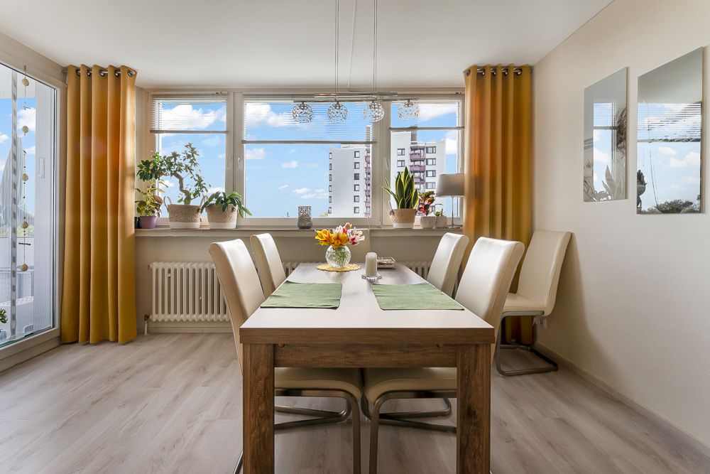 Heller Essbereich mit Holztisch, sechs Stühlen, gelben Vorhängen und vielen Pflanzen am Fenster.