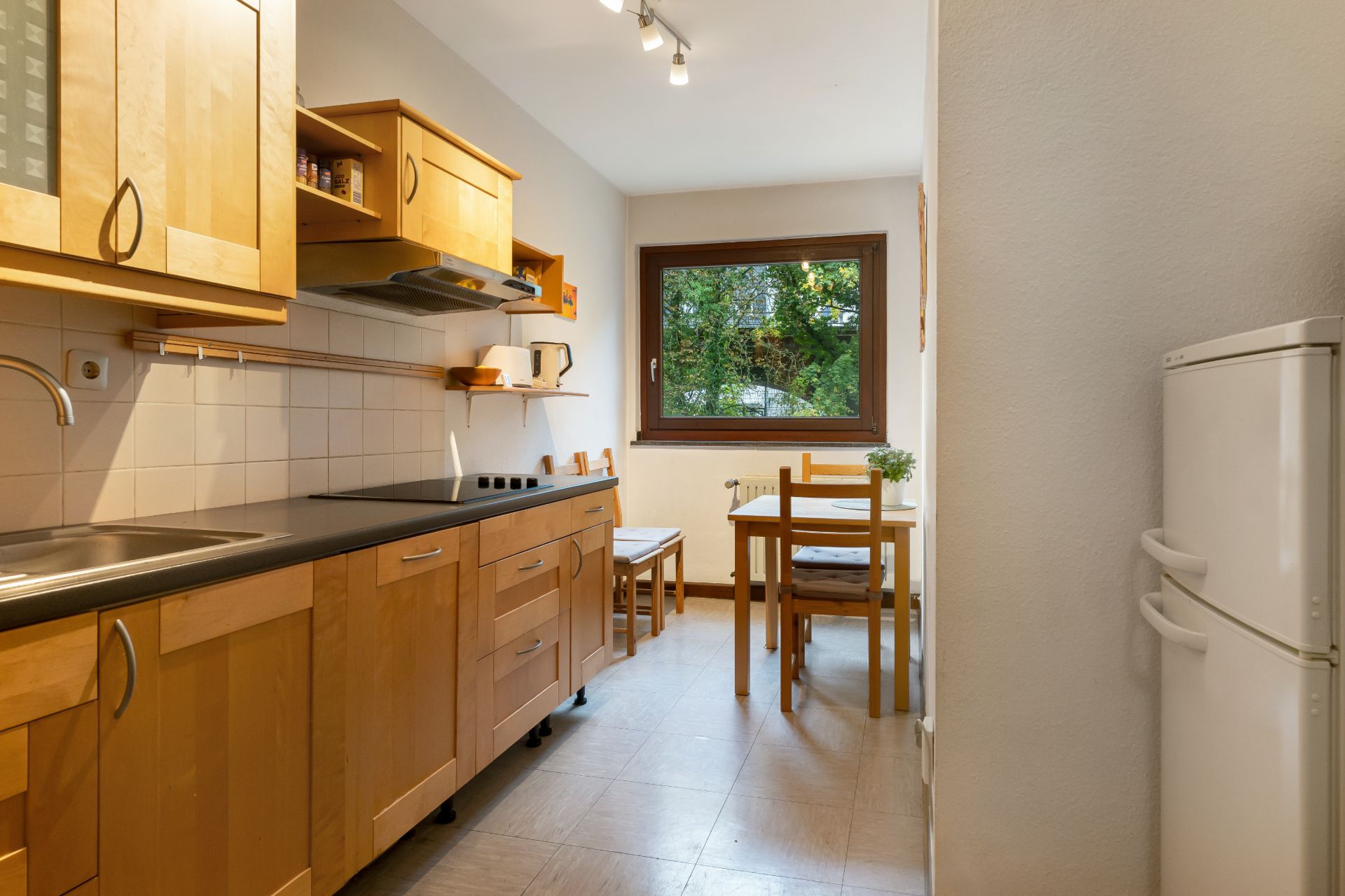 Helle Küche mit Holzschränken, Tisch und zwei Stühlen vor einem Fenster mit Blick ins Grüne.
