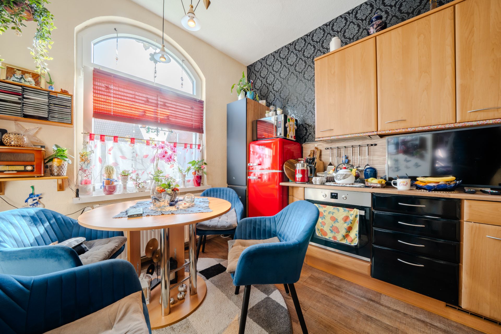 Gemütliche Küche mit Holzmöbeln, rotem Kühlschrank, rundem Tisch und blauen Stühlen am Fenster.