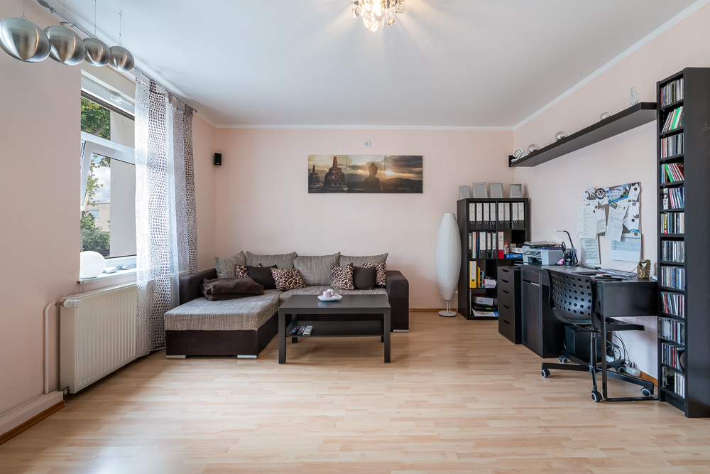 Helles Wohnzimmer mit Ecksofa, Couchtisch, Schreibtisch und Regal auf hellem Holzfußboden.