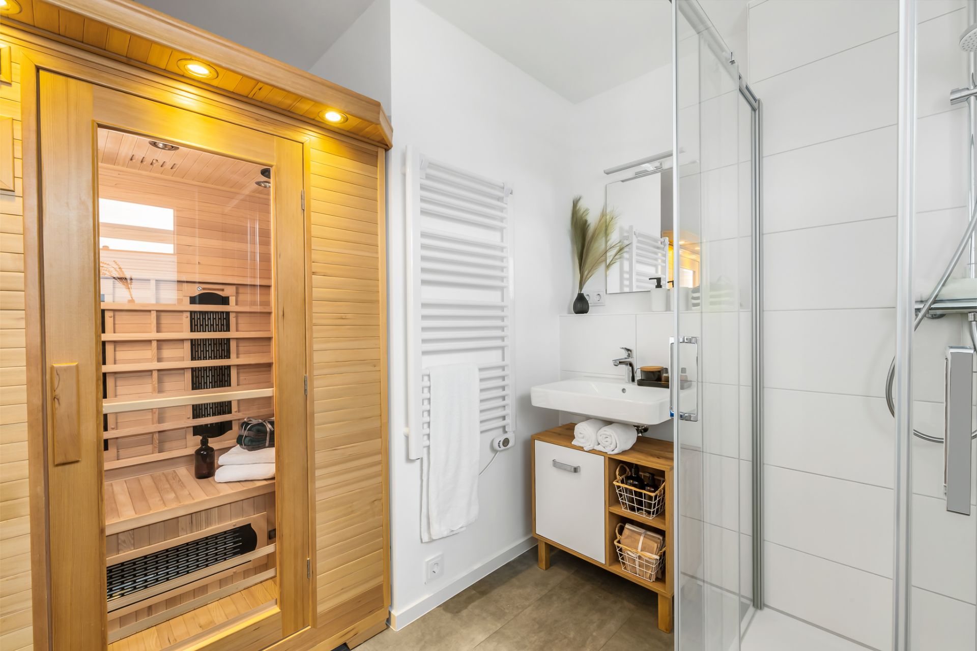 Modernes Badezimmer mit Sauna, Dusche, Waschbecken, Handtuchheizkörper und kleinem Spiegel.