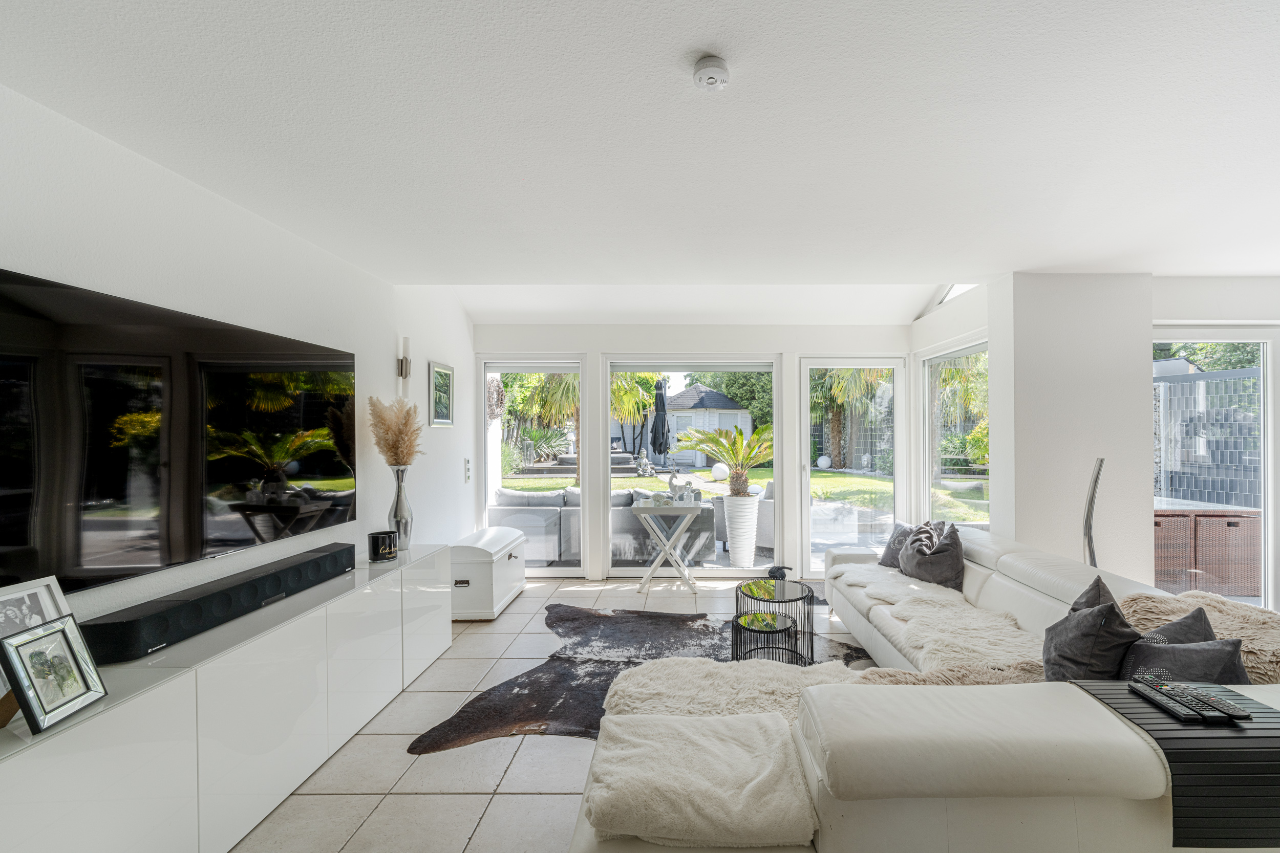 Helles Wohnzimmer mit weißem Sofa, Fernseher, großen Fenstern und Blick in den Garten.