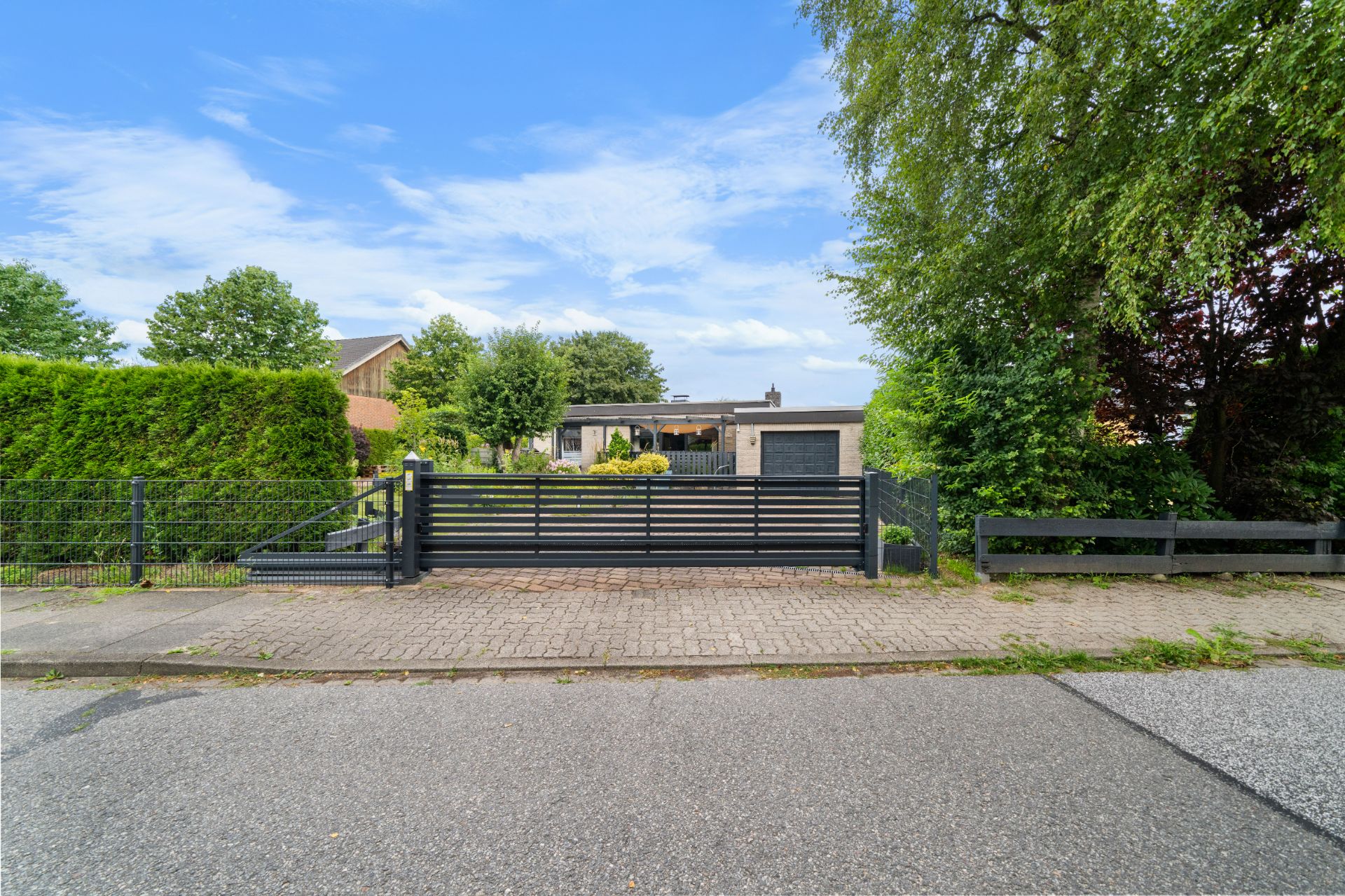 Einfamilienhaus mit Garten, schwarzem Zaun und Tor an ruhiger Straße, umgeben von Bäumen.