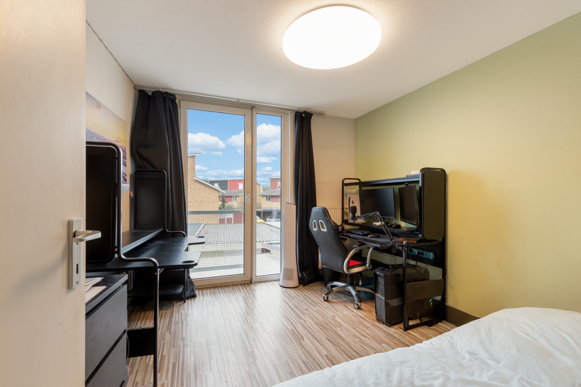 Modernes Schlafzimmer mit Schreibtisch, Stuhl und zwei Monitoren an einem Fenster mit Vorhängen.