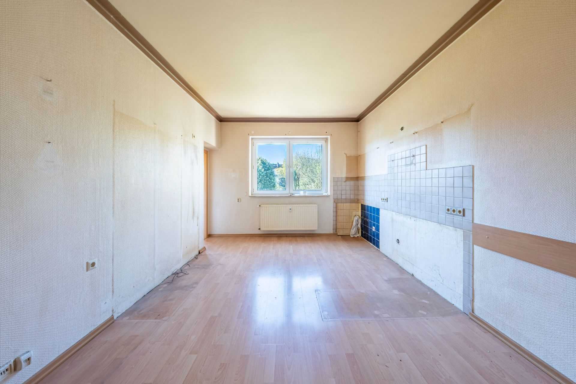 Leerer Raum mit Laminatboden, weißen Wänden und einem Fenster in der Mitte der Rückwand.