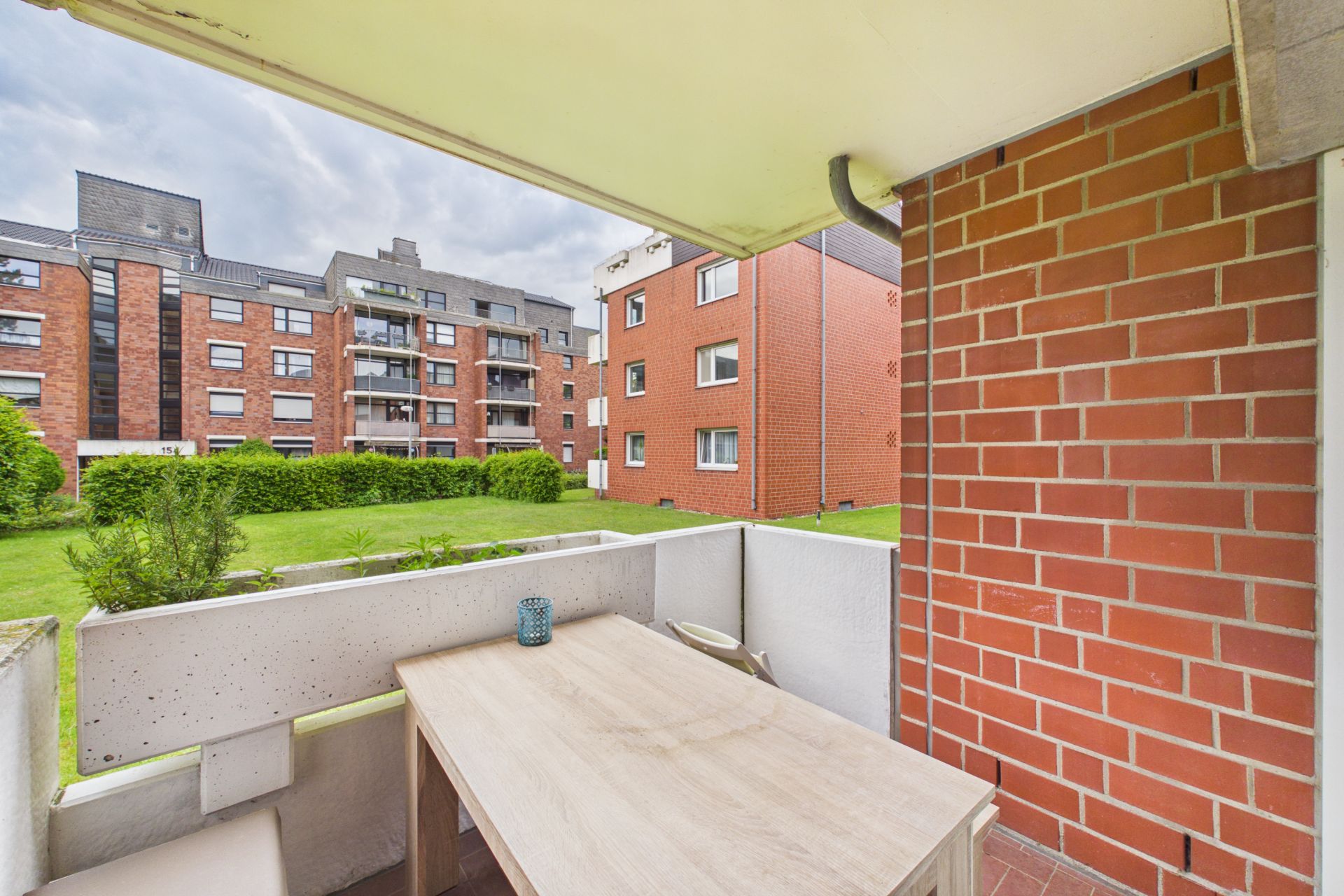 Balkon mit Holztisch und Kerze, Blick auf Wiese und rote Wohngebäude im Hintergrund.