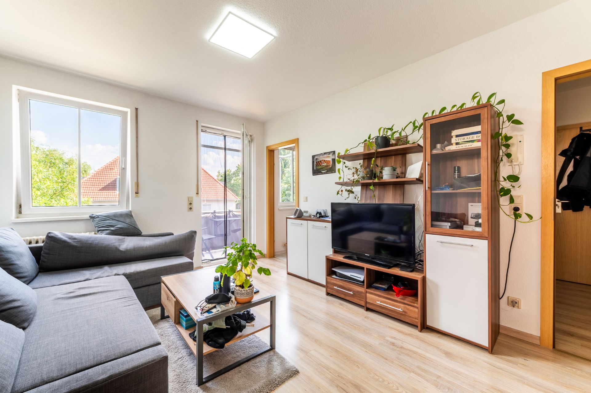Helles Wohnzimmer mit grauem Sofa, Fernseher, Regal, Couchtisch und Balkonzugang.