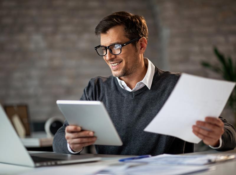 Mann mit Brille arbeitet am Schreibtisch mit Laptop, Tablet und Dokumenten.